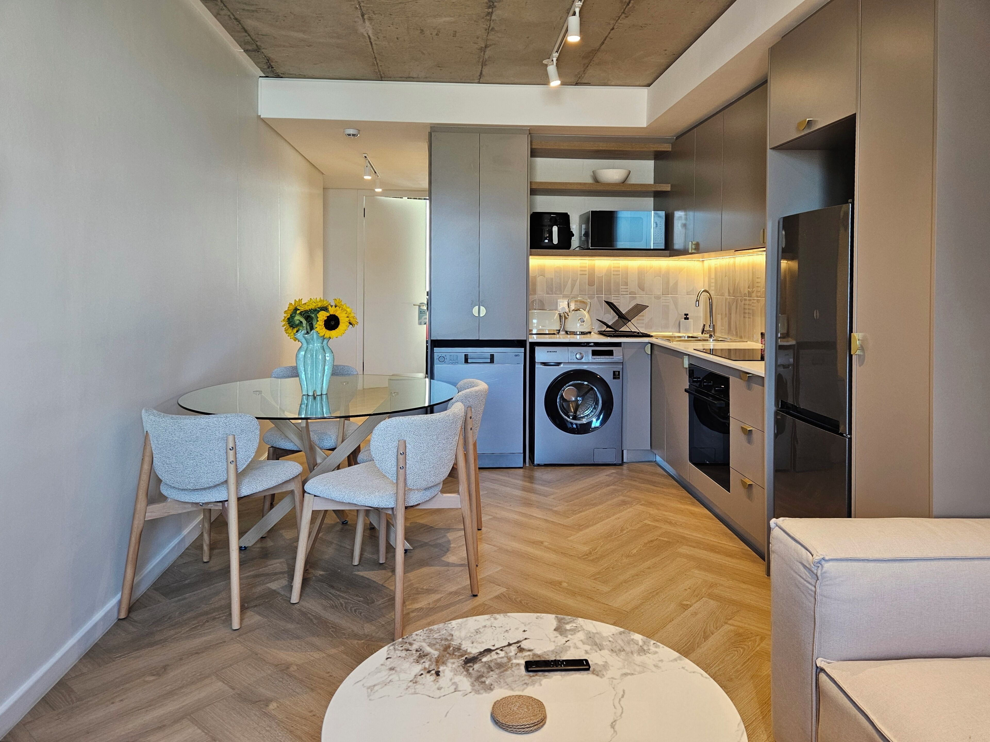 A compact kitchen with modern appliances, a dining area with a glass table and chairs, and a small seating area with a round table.