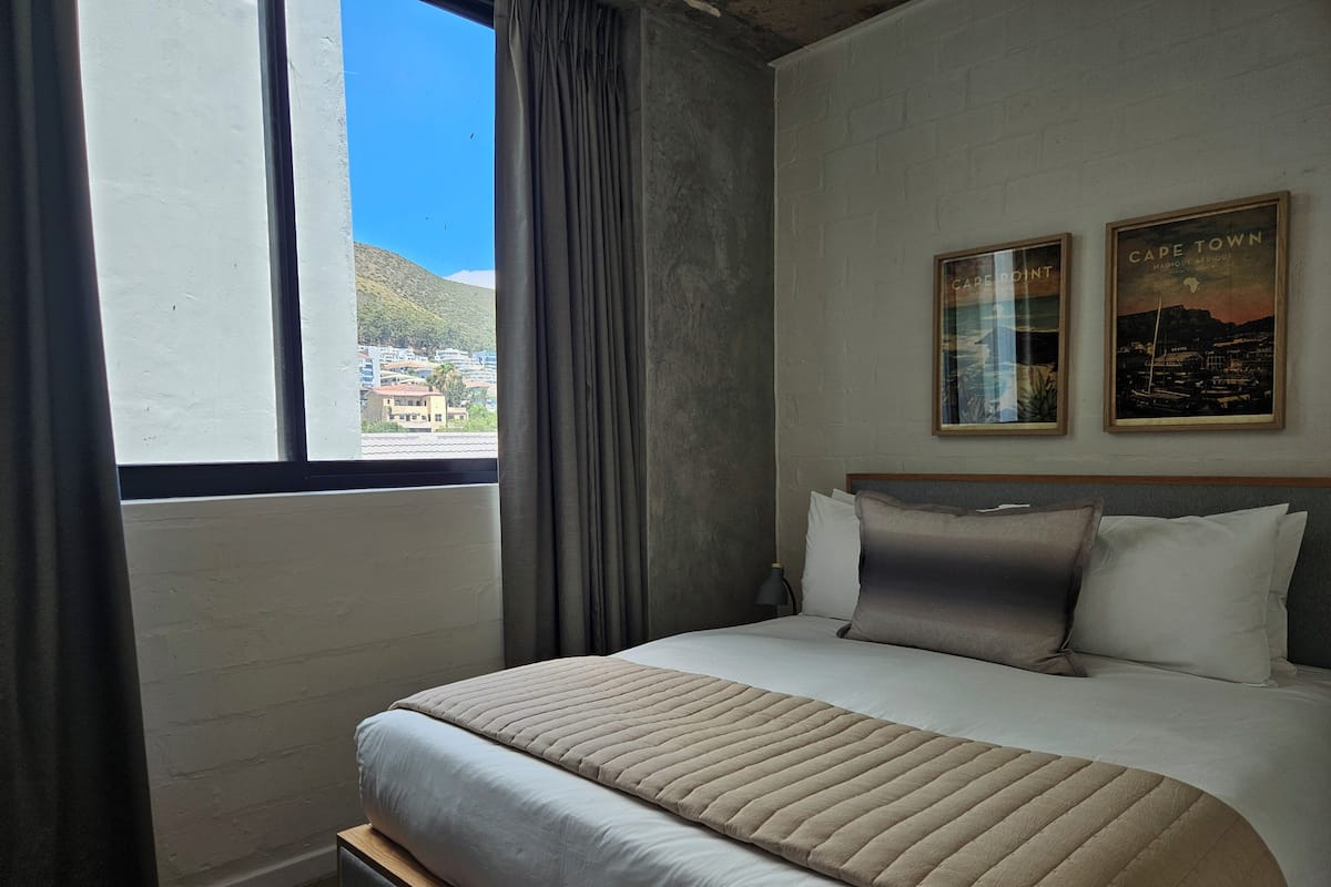 A hotel room with a large bed, a view of a cityscape through a window, and two framed pictures on the wall.