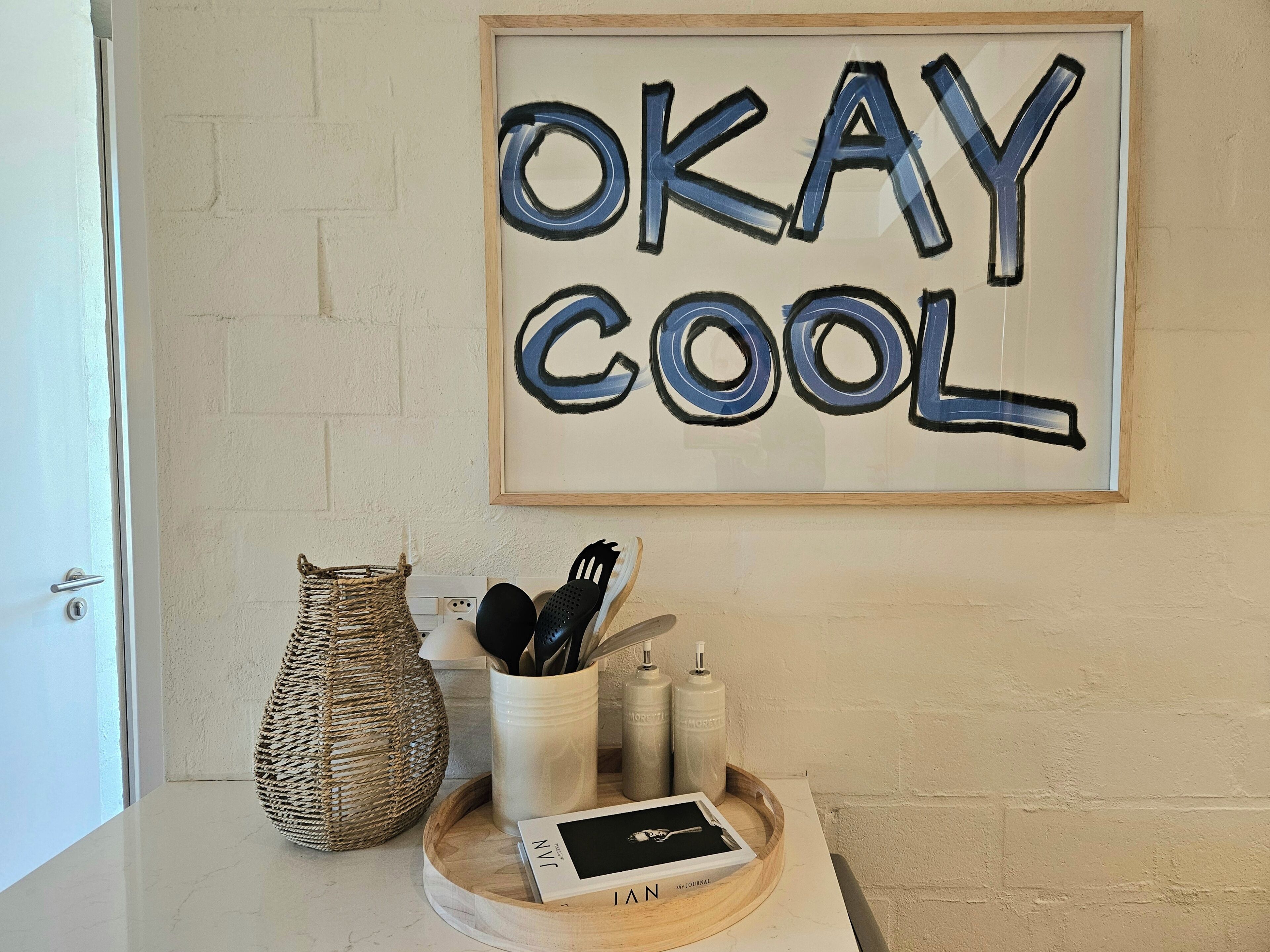 Kehyksiin asetettu taideteos, jossa on valkoisella seinällä teksti ”OKAY COOL”. Sen alla olevalla alustalla on valkoinen muki, musta esine ja pieni valkoinen rasia.
