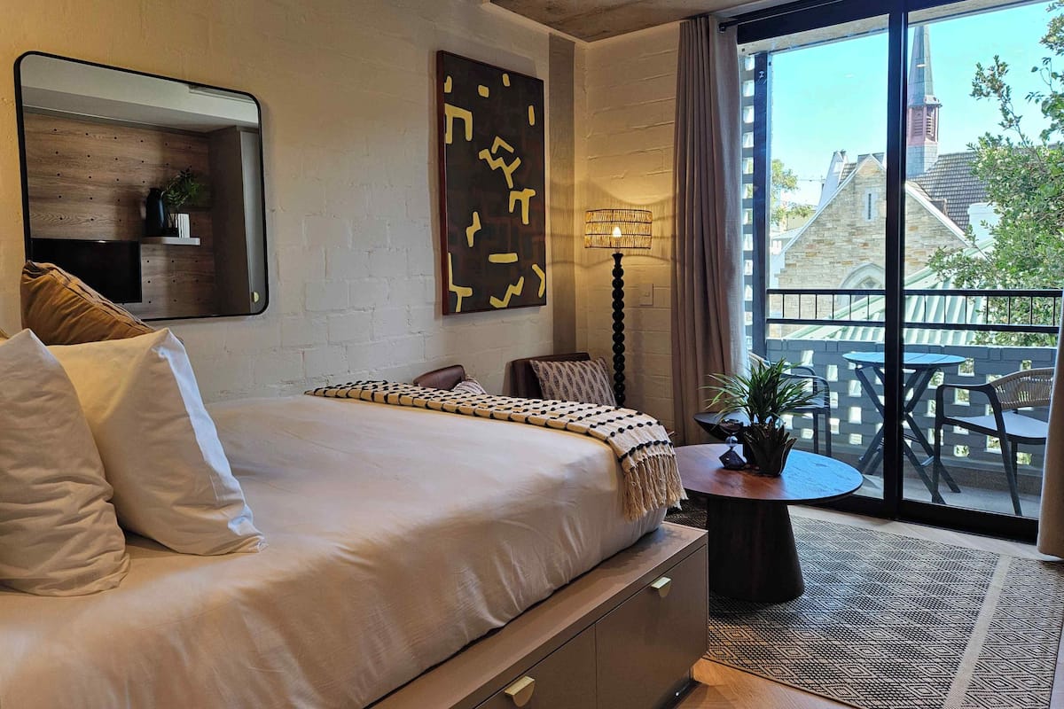 A modern bedroom with a large bed, a side table with a lamp, a mirror, and a view of the outside balcony.
