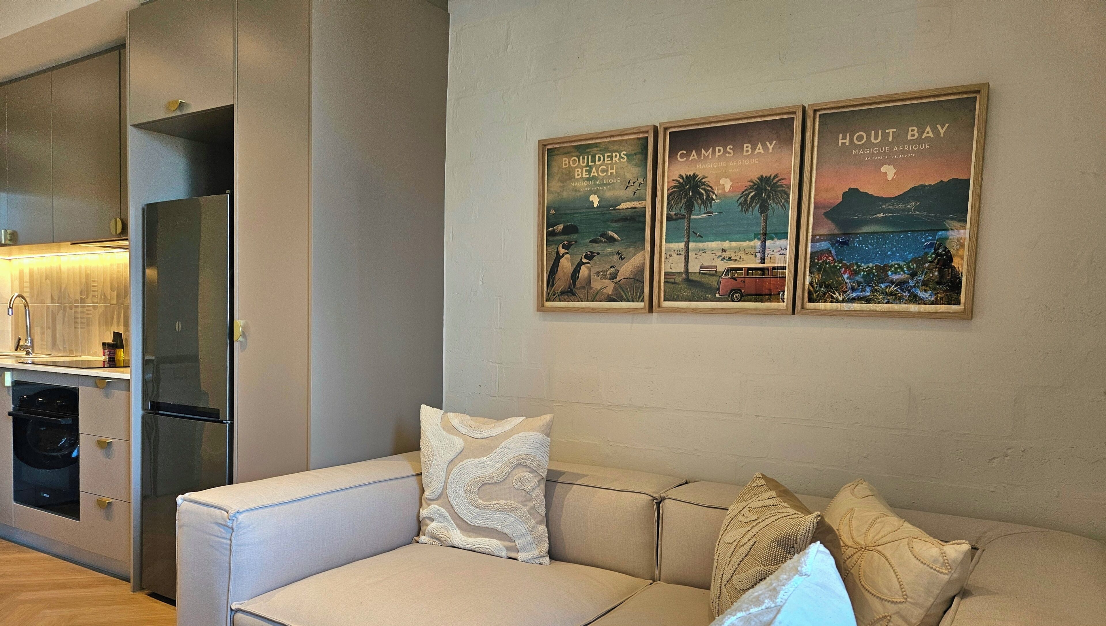 Una sala de estar moderna con un sofá, una zona de cocina con refrigerador, y tres carteles enmarcados en la pared.