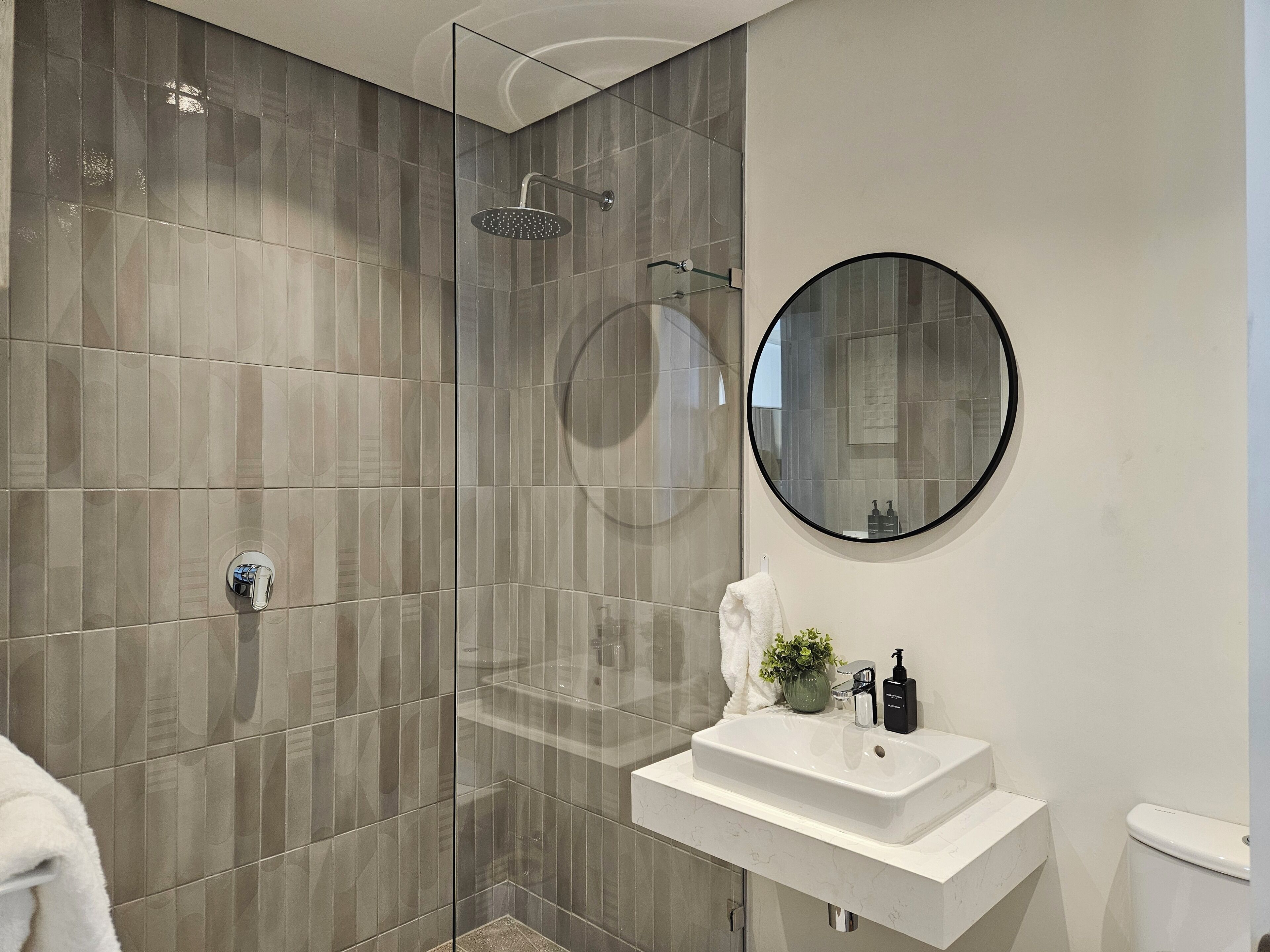 A modern bathroom with a glass shower enclosure, a round mirror, a white sink, and a toilet.