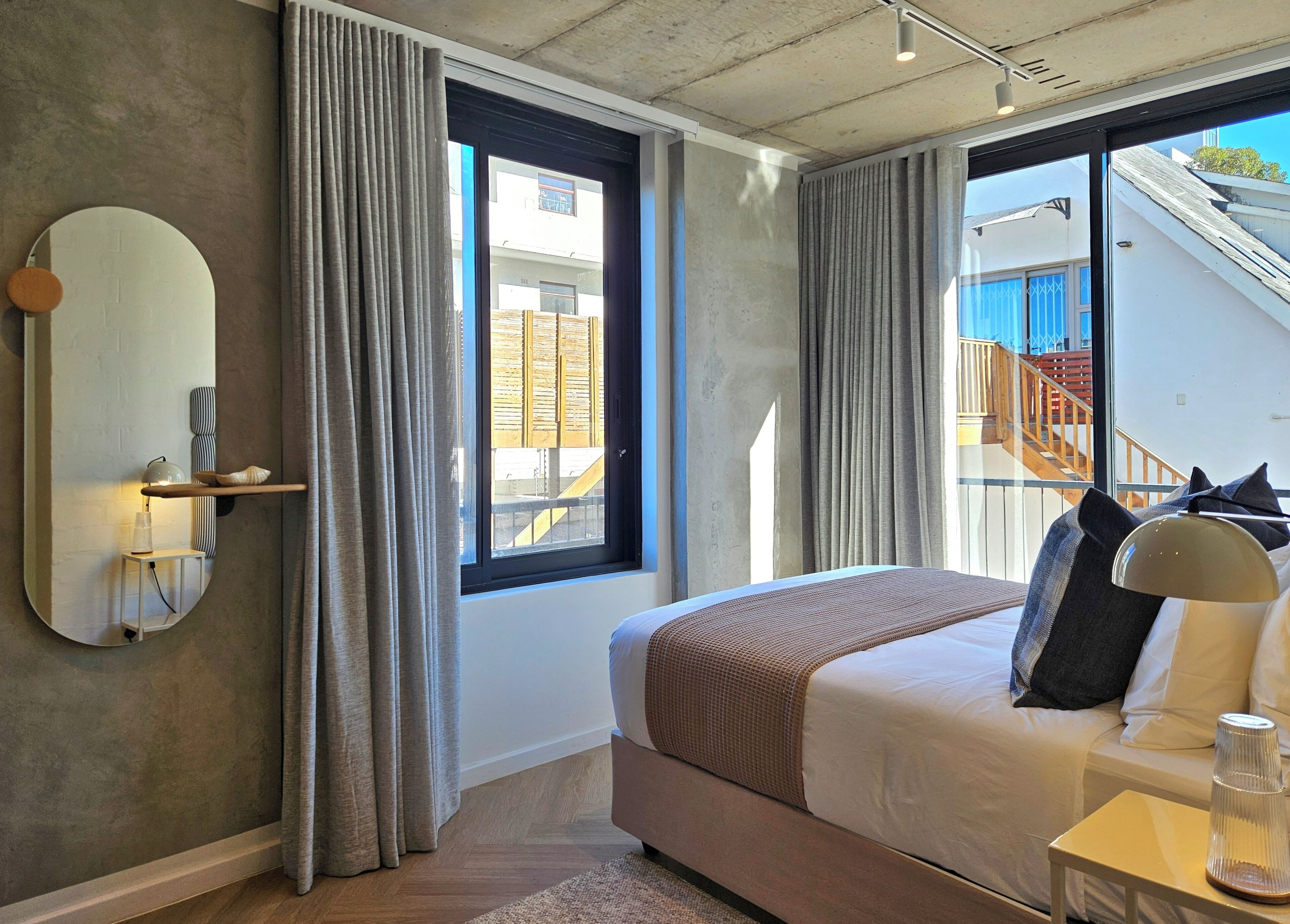 Une chambre d’hôtel moderne avec un grand lit, une table de chevet, un miroir et une vue sur un immeuble avec des escaliers.