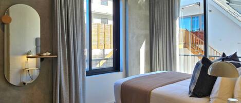 A modern hotel room with a large bed, a bedside table, a mirror, and a view of a building with stairs.