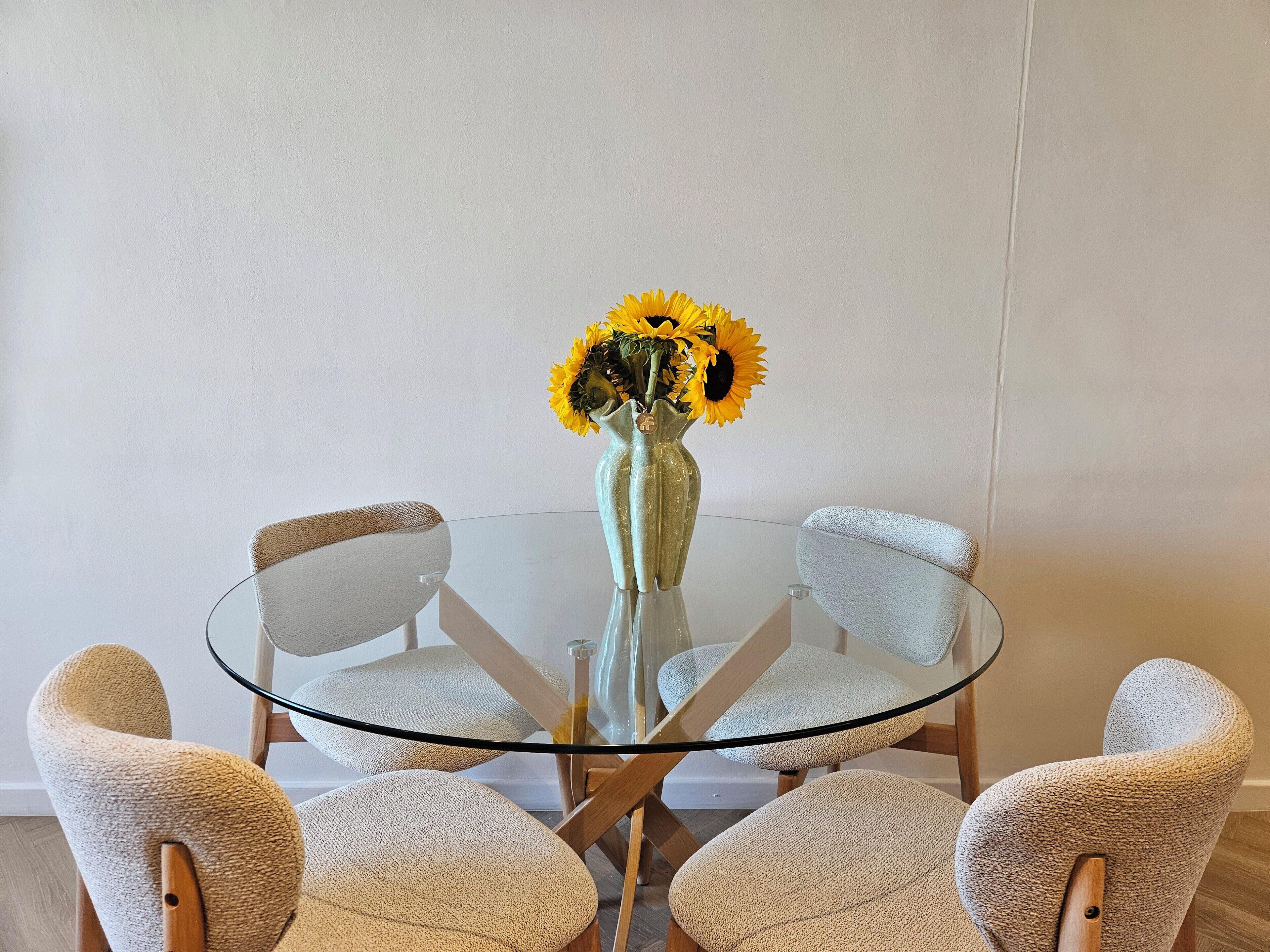 A round glass table with a vase of sunflowers, surrounded by four textured chairs.