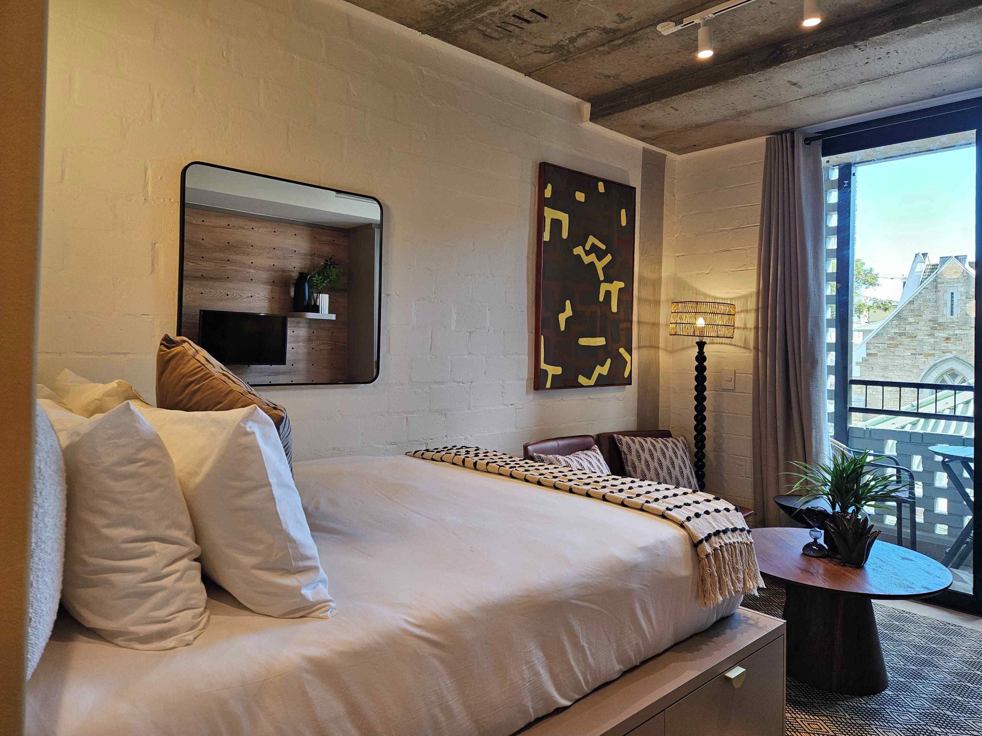 A modern hotel room with a large bed, a small bedside table, a mirror, a wall-mounted shelf with a plant, and a view of the city through large windows.