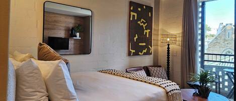 A modern hotel room with a large bed, a small bedside table, a mirror, a wall-mounted shelf with a plant, and a view of the city through large windows.