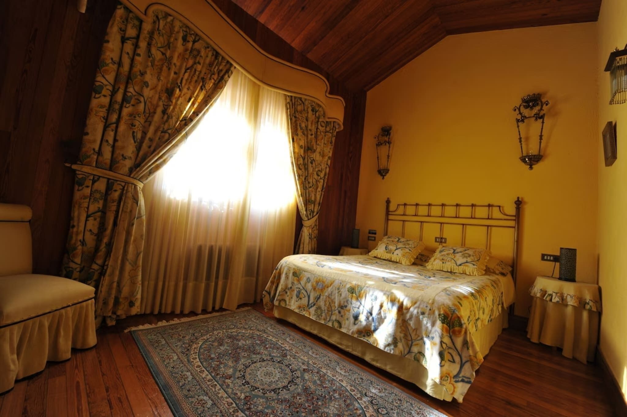 A bedroom with a bed, curtains, a chair, a nightstand, and a wall-mounted lamp.