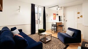 A modern living room with a sofa, armchair, and a coffee table. There is a kitchen area with bar stools and a dining table. The room has white walls and a large window with curtains.