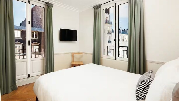 A hotel room with a bed, a TV mounted on the wall, and large windows with curtains.