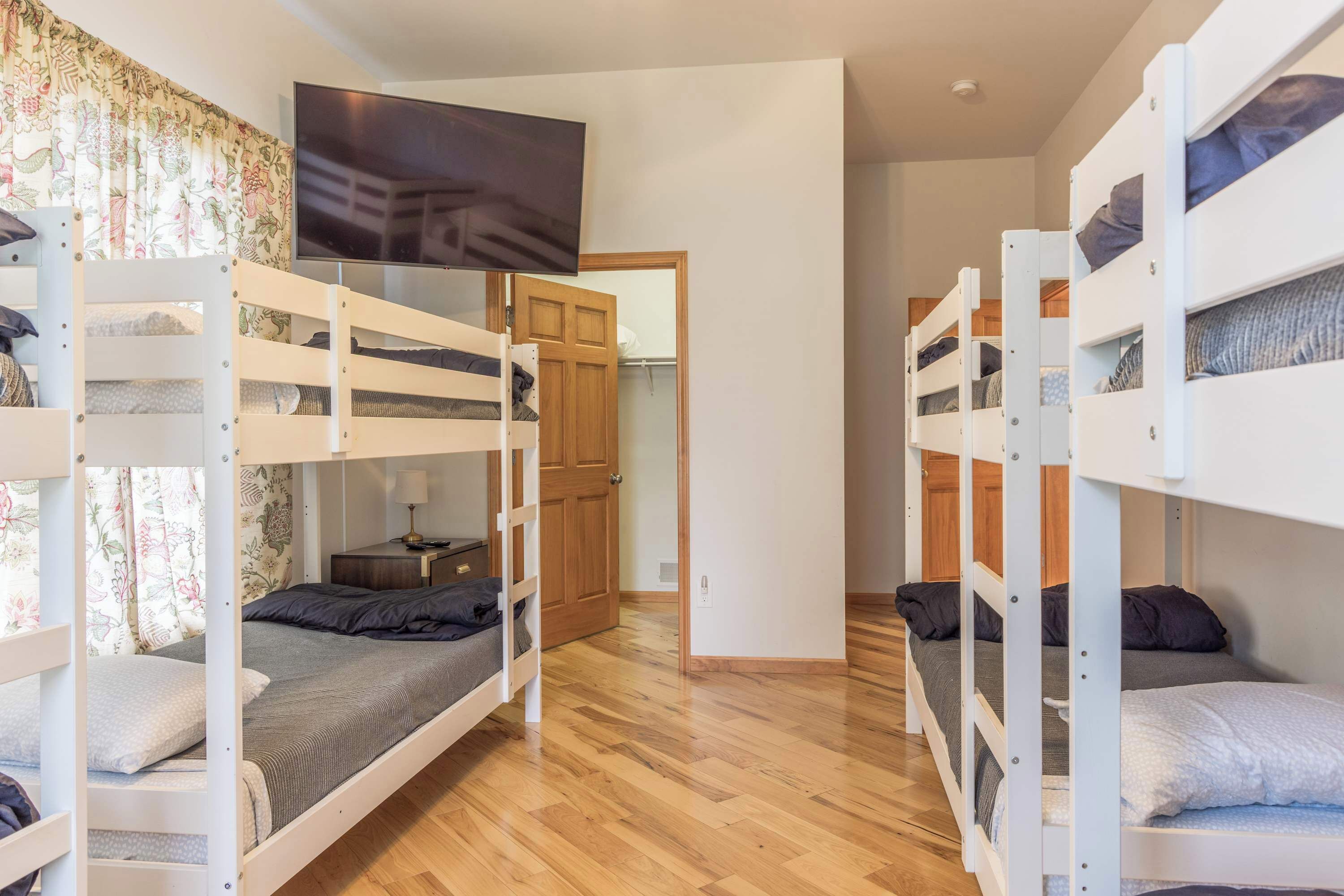 A dormitory room with bunk beds, a television, wooden flooring, and a door leading to another room.