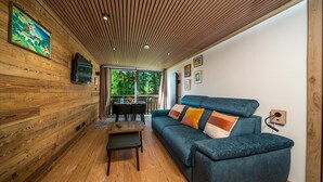 A cozy room with wooden walls, a blue sofa with cushions, a small table, and a TV mounted on the wall. - Beautifully renovated studio apartment, sleeps 4, mountain view (Allos)
