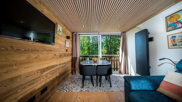 A cozy cabin interior with wooden walls, a dining area with a table and chairs, and a large window offering a view of the forest. - Beautifully renovated studio apartment, sleeps 4, mountain view (Allos)