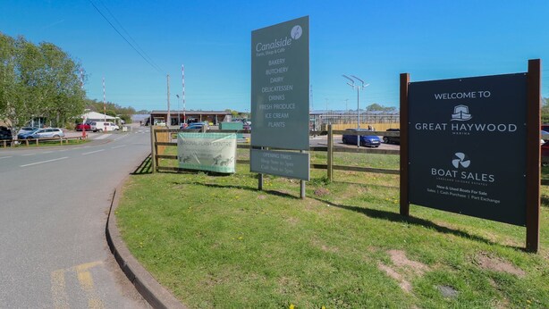 A roadside sign for a seasonal plant center with opening times and a welcome sign for Great Haywood Marina.