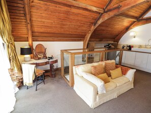 A cozy room with a wooden ceiling, a sofa, a table with a lamp, a mirror, and a view of a kitchen area.