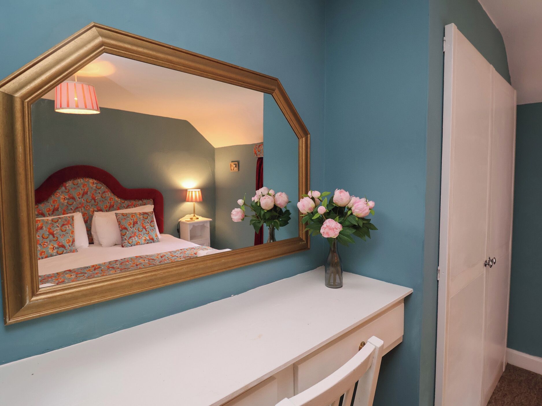 Ein Schlafzimmer mit einem großen Spiegel, einem Bett mit gemusterten Betttextilien, einem Nachttisch mit Lampe und einer Vase mit Blumen.