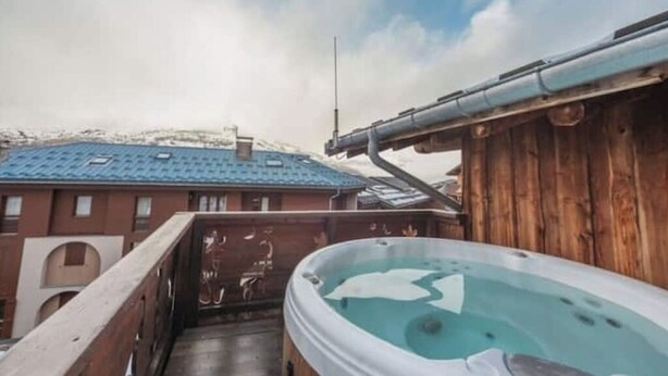 Une terrasse en bois avec un jacuzzi, une rambarde en métal et, au fond, un bâtiment avec un toit bleu.