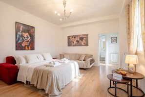 A hotel room with a bed, two sofas, a red chair, a table with a lamp, and artwork on the walls.