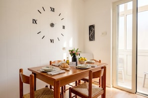 A dining room with a wooden table, four chairs, a wall clock, and a vase with flowers.