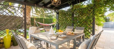 An outdoor patio with a table set for dining, surrounded by chairs, a hammock, and potted plants.