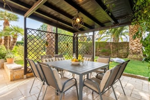 An outdoor patio with a table and chairs, surrounded by a fence, and a view of a garden.