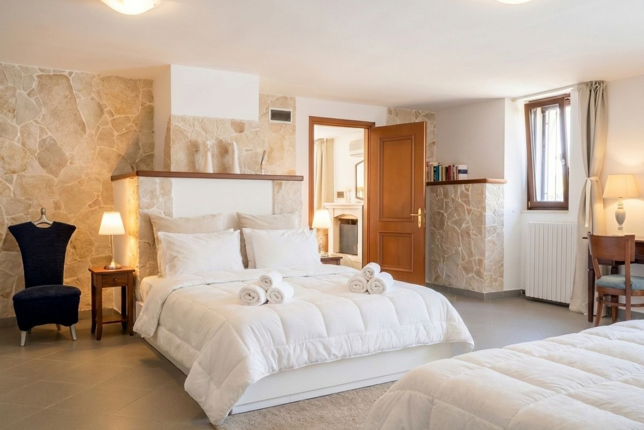 A bedroom with a stone wall, a bed with white bedding, a chair, a lamp, and a wooden door.