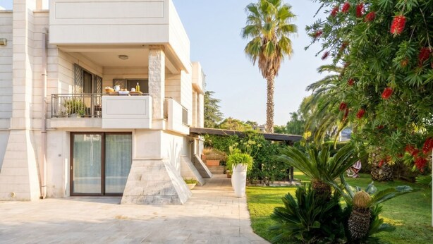 A modern two-story building with a balcony, a paved entrance, and tropical plants.