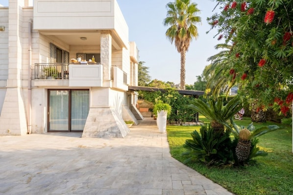 A modern two-story building with a balcony, a paved entrance, and tropical plants.