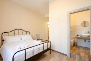 A bedroom with a metal bed frame, white bedding, a bedside table, and a bathroom with a sink and mirror. - Covi del Viaggiatore 2 – Modern Comfort, Timeless Charm (Bari)