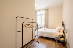 Quarto com cama, cadeira, mesa de cabeceira, radiador e janela com cortinas.