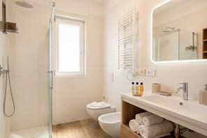 A modern bathroom with a glass-enclosed shower, a toilet, a bidet, a sink with toiletries, and a mirror.