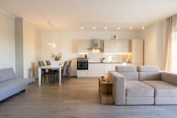A modern living room with a sofa, a dining table with chairs, a kitchen area with cabinets and a stove, and a pendant light.