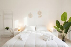 A neatly made bed with white linens, two bedside lamps, a small plant, and a wall-mounted shelf.