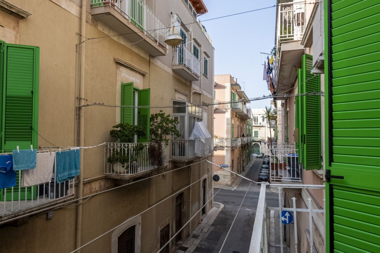 Une rue étroite, bordée de bâtiments des deux côtés, avec des balcons ornés de volets verts et un panneau de signalisation bleu.