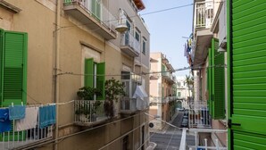 A narrow street with buildings on either side, featuring balconies with green shutters and a blue directional sign.