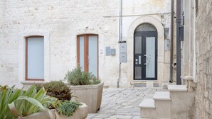 Une cour étroite, pavée de pierres, avec un bâtiment blanc doté de deux portes : l’une avec un cadre en bois et l’autre avec une porte noire. On aperçoit des plantes en pot au premier plan.