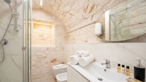 Un baño con ducha de vidrio, un lavamanos blanco con artículos de aseo y una pared de piedra.