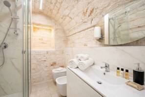 Un baño con ducha de vidrio, un lavamanos blanco con artículos de aseo y una pared de piedra.