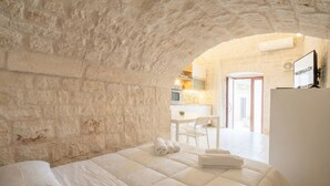Un dormitorio con pared de piedra, cama con ropa de cama blanca, y vista a una cocina y comedor a través de un arco.