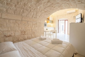 Un dormitorio con pared de piedra, cama con ropa de cama blanca, y vista a una cocina y comedor a través de un arco.