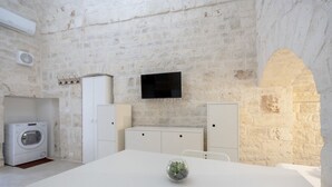 Une pièce avec un mur en pierre, un téléviseur à écran plat fixé au-dessus d’un meuble blanc, un lave-linge et une table à manger agrémentée d’un vase en verre.