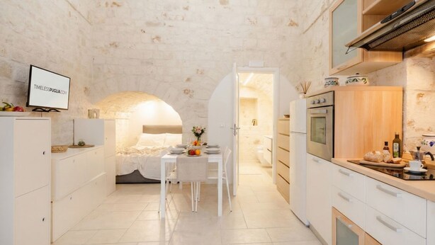 Cocina compacta con muebles blancos, una zona de comedor con mesa para dos personas y un horno a la vista.