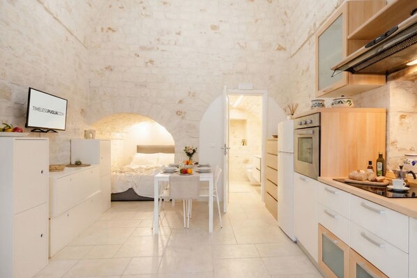 Cocina compacta con muebles blancos, una zona de comedor con mesa para dos personas y un horno a la vista.