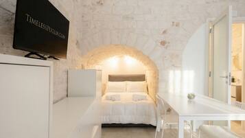 Una habitación con pared de piedra, una cama con ropa de cama blanca, un televisor montado en la pared y una mesa blanca con sillas.