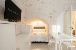 Una habitación con pared de piedra, una cama con ropa de cama blanca, un televisor montado en la pared y una mesa blanca con sillas.