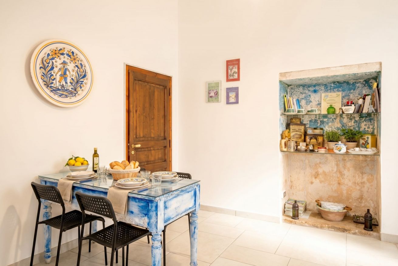 Un comedor con una mesa dispuesta para cuatro personas, una puerta de madera y una estantería con diversos objetos.