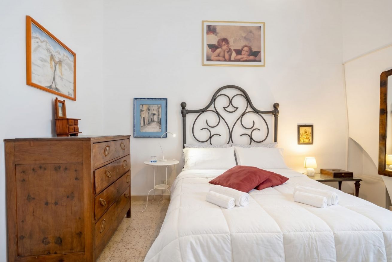 A bedroom with a wooden dresser, a bed with white bedding and a red pillow, a nightstand with a lamp, and a wall with framed pictures.