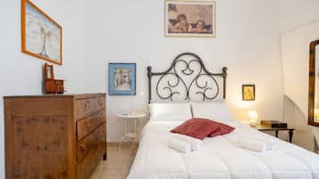 A bedroom with a wooden dresser, a bed with white bedding and a red pillow, a nightstand with a lamp, and a wall with framed pictures.