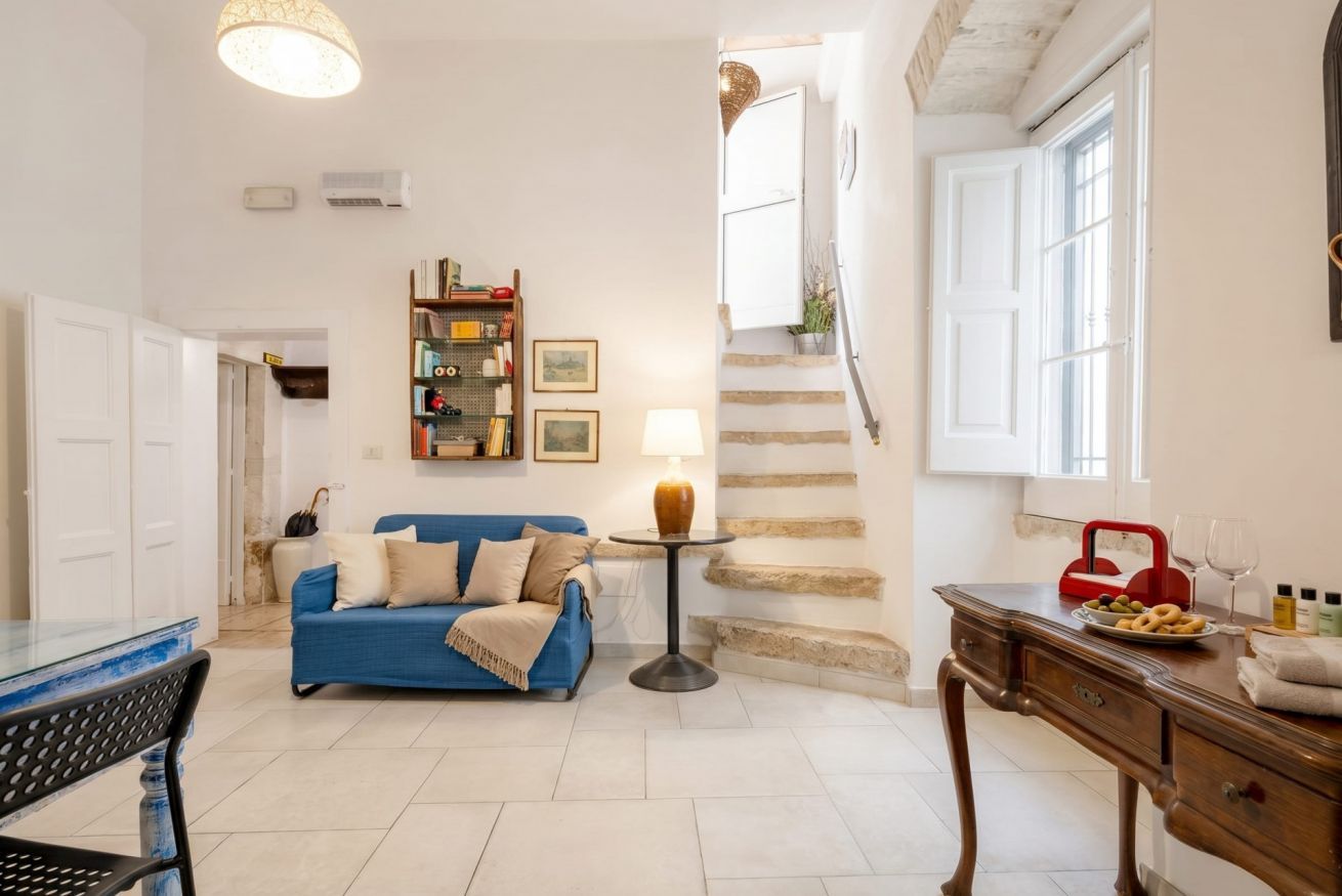 Una sala de estar con un sofá azul, una mesa de centro, una lámpara, una estantería y una escalera.