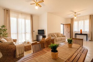 A spacious living area with a dining table, wicker furniture, a television, and a ceiling fan.
